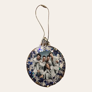 May include: A round Christmas ornament with a photo of five people in light-colored suits. The ornament is decorated with blue and silver glitter and has a gold string for hanging. The ornament is a festive holiday decoration.