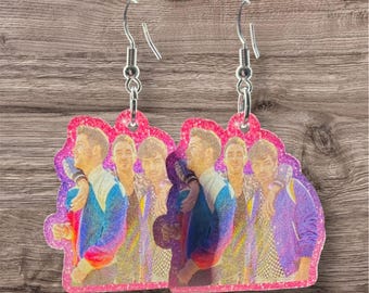 May include: A pair of glittery dangle earrings featuring a group of people in colorful jackets. The earrings have a pink border and silver-colored fish hook ear wires. The image on the earrings has a purple and pink background.
