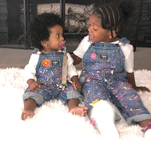 Puede incluir: Dos niños pequeños con monos de mezclilla con salpicaduras de pintura de colores. El niño de la izquierda lleva una camisa blanca y un chupete. El niño de la derecha lleva una camiseta blanca. Ambos niños están sentados en una alfombra blanca y esponjosa.