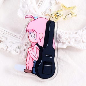 May include: A cartoon keychain featuring a pink-haired character with a worried expression, leaning against a black guitar case. The keychain has a gold star-shaped clasp. The character is wearing a pink outfit.