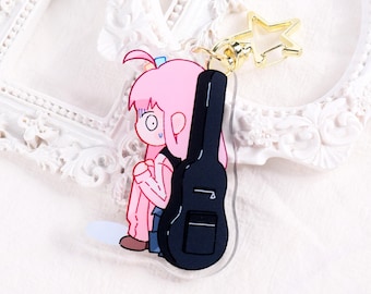 May include: A cartoon keychain featuring a pink-haired character with a worried expression, leaning against a black guitar case. The keychain has a gold star-shaped clasp. The character is wearing a pink outfit.