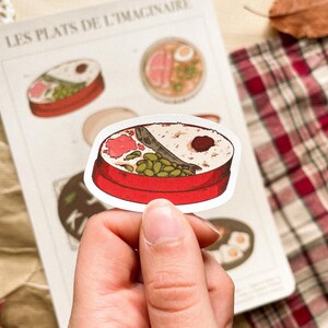 May include: A sticker featuring a red bento box with rice, fish, and vegetables. The sticker is held by a hand, with other food illustrations visible on a paper background. The text "LES PLATS DE L'IMAGINAIRE" is at the top.