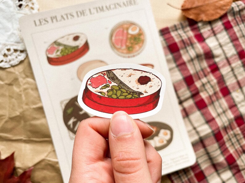 May include: A sticker featuring a red bento box with rice, fish, and vegetables. The sticker is held by a hand, with other food illustrations visible on a paper background. The text "LES PLATS DE L'IMAGINAIRE" is at the top.