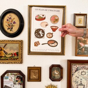 May include: A collection of framed art pieces on a white wall. The art includes a framed print with the text "LES PLATS DE L'IMAGINAIRE", an oval floral embroidery, a windmill tapestry, and other small framed pieces.