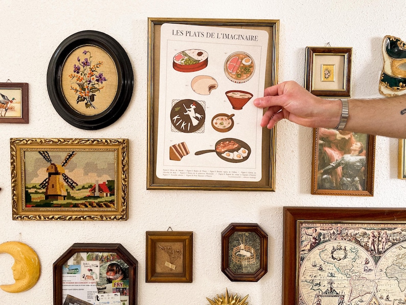 May include: A collection of framed art pieces on a white wall. The art includes a framed print with the text "LES PLATS DE L'IMAGINAIRE", an oval floral embroidery, a windmill tapestry, and other small framed pieces.