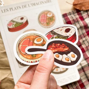 May include: A hand holding a collection of food-themed stickers. The stickers feature illustrations of various dishes, including ramen, sushi, and fried eggs. The background includes a page with the text "LES PLATS DE L'IMAGINAIRE".
