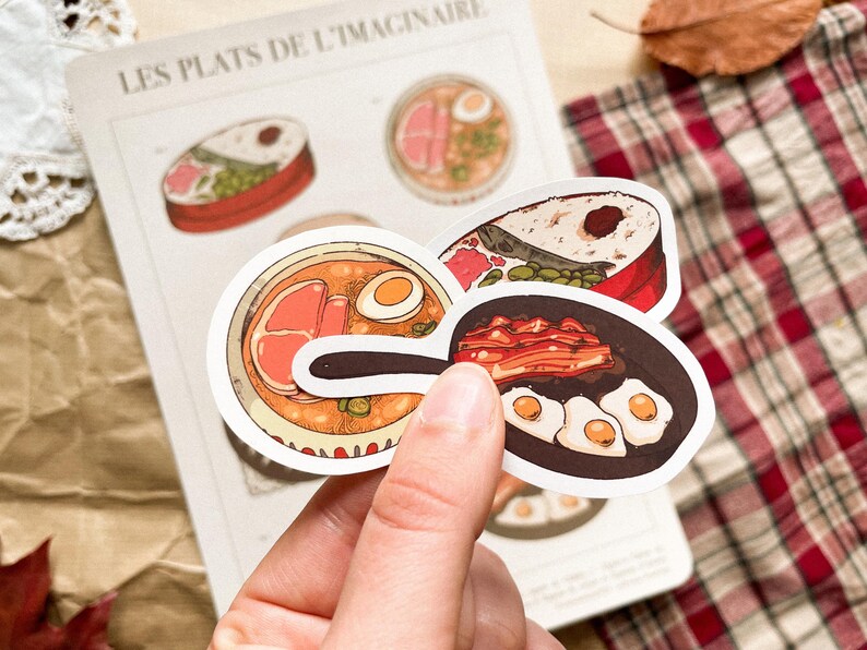 May include: A hand holding a collection of food-themed stickers. The stickers feature illustrations of various dishes, including ramen, sushi, and fried eggs. The background includes a page with the text "LES PLATS DE L'IMAGINAIRE".