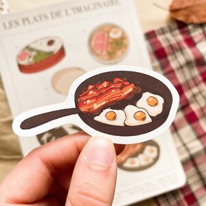 May include: A sticker featuring a cartoon illustration of a skillet with fried eggs and bacon. The sticker is white with a black handle and a brown skillet. The eggs have yellow yolks. The sticker is held in a person's hand.