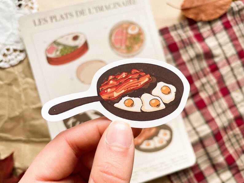 May include: A sticker featuring a cartoon illustration of a skillet with fried eggs and bacon. The sticker is white with a black handle and a brown skillet. The eggs have yellow yolks. The sticker is held in a person's hand.