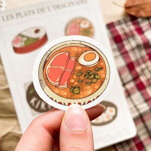 May include: A round sticker featuring a bowl of ramen with a soft-boiled egg, meat, and green onions. The sticker is held by a hand, with a notepad and plaid fabric in the background. The sticker is part of a series called "Les Plats de l'Imaginaire."