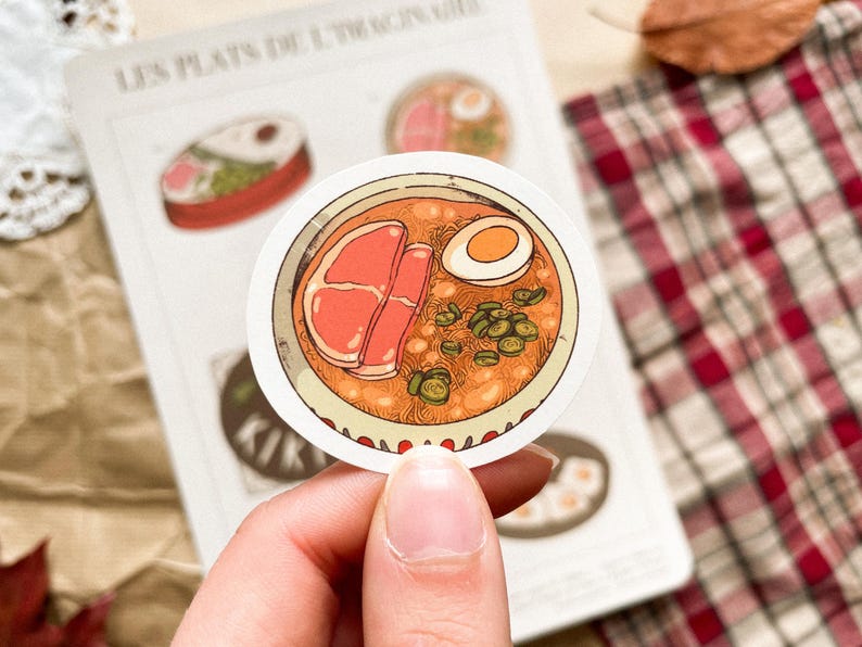 May include: A round sticker featuring a bowl of ramen with a soft-boiled egg, meat, and green onions. The sticker is held by a hand, with a notepad and plaid fabric in the background. The sticker is part of a series called "Les Plats de l'Imaginaire."