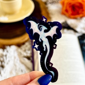 May include: A die-cut sticker featuring a light blue, slender dragon with outstretched wings against a dark navy blue background. The design includes small white star shapes scattered around the dragon, which has a long, curved tail.