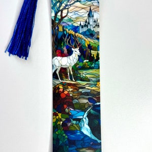 May include: A rectangular bookmark featuring a stained glass style illustration of a white stag standing in a forest landscape with a castle in the background. The bookmark is topped with a vibrant blue tassel attached by a thin blue cord.