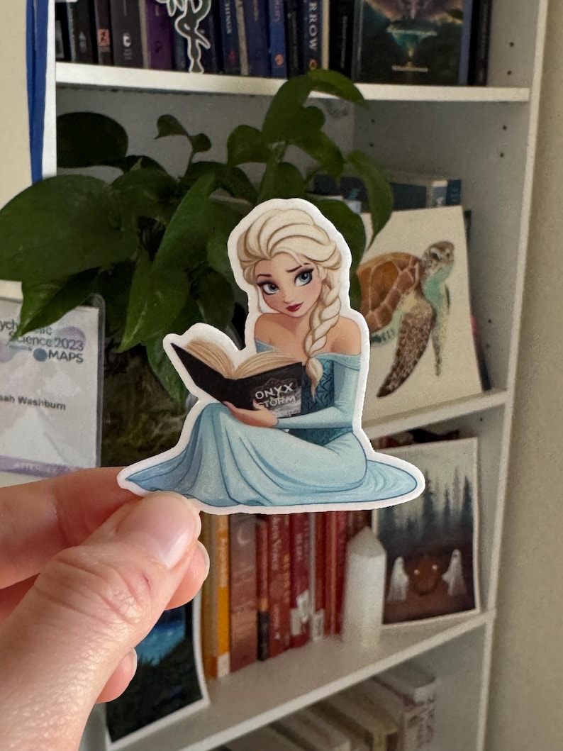 May include: A sticker featuring a cartoon illustration of a young woman with long blonde hair, wearing a blue dress and reading a book titled "Onyx Storm".