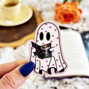 May include: A die-cut sticker featuring a white ghost character covered in small pink hearts. The ghost has two black oval eyes and is holding an open black book with a detailed illustration on the cover. The sticker has a thin pink border.