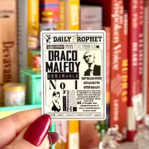 Könnte beinhalten: Ein rechteckiger Aufkleber mit einem Zeitungsdesign, der mit "DAILY PROPHET" überschrieben ist. Der Aufkleber zeigt "DRACO MALFOY DESIRABLE No 1" in fetter Schrift, zusammen mit Schwarz-Weiß-Porträts und Text, der einem Zeitungsartikel ähnelt.