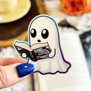 May include: A die-cut sticker featuring a white ghost with large black eyes holding an open book. The book cover is black with white text that reads When The Moon Hatched by Sarah J. Parker. The sticker has a thin blue and purple border.