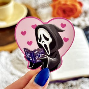 May include: A heart-shaped sticker featuring a cartoon figure in a black hooded robe with a white ghost mask, reading a purple book titled Assistant to the Villain. The background of the sticker is light pink with several small dark pink hearts.