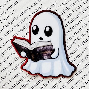 May include: A white ghost sticker with large black eyes holding an open book. The book cover is black with the text ALCHEMIST printed in gold. The sticker has a thin red border and is placed on a page of printed text with black ink.