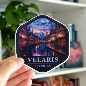 May include: A sticker featuring a nighttime scene of a town with a river running through it. The town is lit up with lights and there are mountains in the background. The sticker is hexagonal and has the text "VELARIS PRYTHIAN" at the bottom.