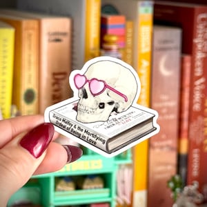May include: A sticker of a skull wearing pink heart-shaped glasses, placed on a book. The book's title reads "Draco Malfoy & the Mortifying Ordeal of Being in Love". The sticker is held in front of a bookshelf.
