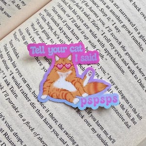 Puede incluir: Una pegatina con un gato atigrado naranja con gafas de sol en forma de corazón. La pegatina tiene el texto "Tell your cat I said pspsps" en rosa y azul. El gato está sobre un fondo lila claro.