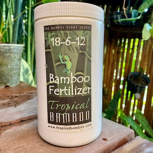 May include: White plastic container of "Bamboo Fertilizer" with a label featuring a frog and bamboo stalks. The label also displays the numbers "18-6-12" and the website address www.tropicalbamboo.com. The container is on a wooden surface.
