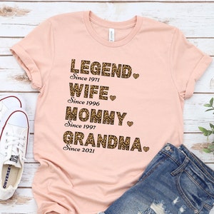May include: A peach-colored t-shirt with a leopard print design. The text on the shirt reads "LEGEND. Since 1971 WIFE. Since 1996 MOMMY. Since 1997 GRANDMA. Since 2021".