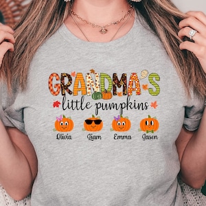May include: A light gray t-shirt with the text "GRANDMA'S little pumpkins" in colorful letters and pumpkin graphics. Below the text are four cartoon pumpkins with names: Olivia, Liam, Emma, and Jason. The shirt is made of Athletic Heather material.