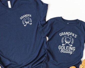 Può includere: Due magliette blu navy con un design a tema golf. La maglietta più grande dice "Grandpa" con una palla da golf e mazze. La maglietta più piccola dice "Grandpa's Golfing Buddy".