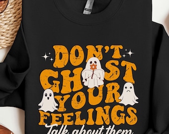 Op de afbeelding: Zwart sweatshirt met de tekst "DON'T GHOST YOUR FEELINGS Talk about them" in oranje, met drie witte spookillustraties. Eén spook houdt een bloem vast. Het sweatshirt heeft een ronde hals.