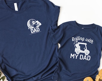 May include: Two navy blue t-shirts with white text and graphics. The larger shirt says "DAD" and has a golf ball and club graphic. The smaller shirt says "Rolling with my DAD" and has a golf cart graphic.