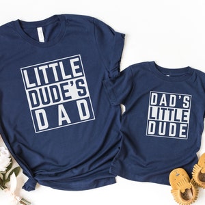 May include: Two navy blue t-shirts with white text. The larger shirt says "Little Dude's Dad" and the smaller shirt says "Dad's Little Dude".