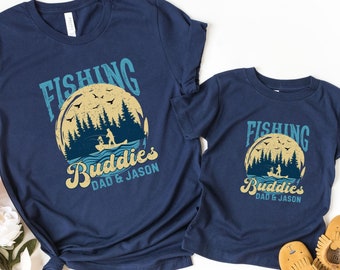 Könnte beinhalten: Zwei marineblaue T-Shirts mit einer Grafik im Vintage-Stil eines Mondes, Bäumen und zwei Personen, die in einem Boot angeln. Der Text auf den T-Shirts lautet "Fishing Buddies Dad & Jason".