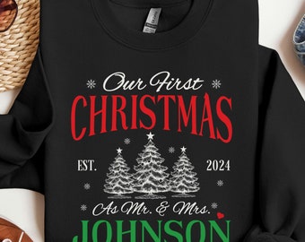 May include: Black sweatshirt with white text that reads "Our First Christmas * EST. 2024 * As Mr. & Mrs. JOHNSON" with a red heart. The text is surrounded by white snowflakes and three white stylized Christmas trees.