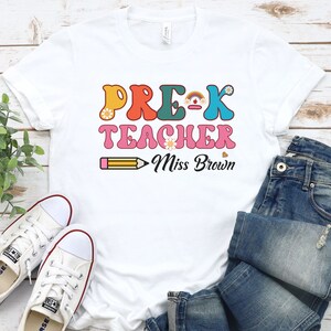 May include: White t-shirt with the text "Pre-K Teacher Miss Brown" in colorful, retro-style letters. The text is decorated with a rainbow, flowers, and a pencil.