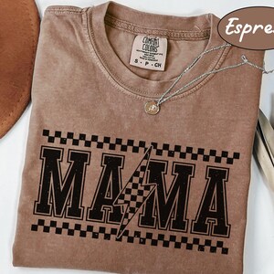 May include: Espresso-colored t-shirt with the word "MAMA" in a black, distressed, collegiate-style font, with a checkered pattern above and a lightning bolt graphic. The shirt is a Comfort Colors brand, and a silver necklace with a pendant is visible.