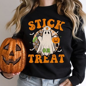 May include: Black sweatshirt with a Halloween graphic. The design shows a ghost in a nurse's cap, holding a syringe and a jack-o'-lantern bucket. The words "STICK OR TREAT" are in orange and green, with bats and a spider web.