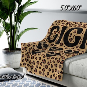 K&ouml;nnte beinhalten: Eine braune und beige Leopardenfell-Fleece-Decke mit dem Wort "BIG" in schwarzen Buchstaben. Die Decke ist auf einem Sofa gefaltet und hat drei Herzen mit Namen darauf. Die Decke ist 50 Zoll mal 60 Zoll gro&szlig;.