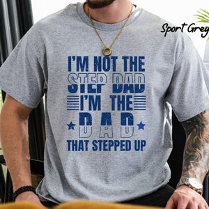 Puede incluir: Una camiseta gris con el texto "I'm not the step dad I'm the dad that stepped up" en letras azules. La camiseta tiene un aspecto desgastado y presenta una estrella a cada lado de la palabra "dad".