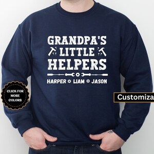 May include: Navy blue sweatshirt with white text that reads "Grandpa's Little Helpers" with a wrench and hammer graphic. Below the text is a line with three wrenches and the names "Harper, Liam, Jason". The text "Customizable" is in the bottom right corner.