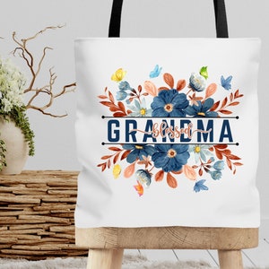 K&ouml;nnte beinhalten: Wei&szlig;e Einkaufstasche mit einem Blumendesign in Blaut&ouml;nen, Orange und Gr&uuml;n. Der Text "blessed grandma" ist in Blau auf die Tasche gedruckt.