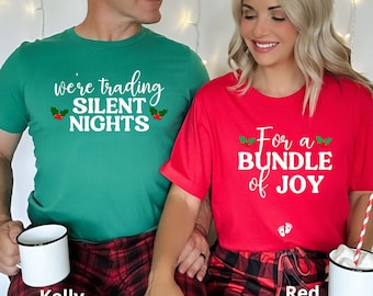 May include: Couple wearing matching Christmas themed shirts. The man's shirt says "We're trading silent nights" with holly sprigs. The woman's shirt says "For a bundle of joy" with holly sprigs and footprints. They are both wearing red and black plaid pajamas.