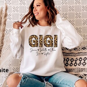May include: White sweatshirt with the word "GIGI" in leopard print letters. Below the word are the names "James", "Isabella", "Olivia", "Liam", and "Sophia" with hearts between each name.