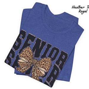 Puede incluir: Una camiseta azul con la palabra "Senior" impresa en negro con un lazo de estampado de leopardo. La camiseta est&aacute; doblada por la mitad.