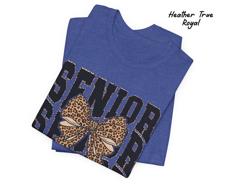 Puede incluir: Una camiseta azul con la palabra "Senior" impresa en negro con un lazo de estampado de leopardo. La camiseta est&aacute; doblada por la mitad.