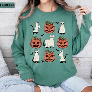 K&ouml;nnte beinhalten: Hellgr&uuml;nes Sweatshirt mit Halloween-Muster. Das Design zeigt orangefarbene K&uuml;rbislaternen, wei&szlig;e Geisterkatzen und schwarze Flederm&auml;use. Die Worte "LIGHT GREEN" stehen in einem gr&uuml;nen Rechteck oben links.