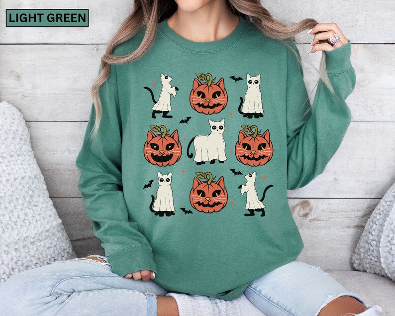 K&ouml;nnte beinhalten: Hellgr&uuml;nes Sweatshirt mit Halloween-Muster. Das Design zeigt orangefarbene K&uuml;rbislaternen, wei&szlig;e Geisterkatzen und schwarze Flederm&auml;use. Die Worte "LIGHT GREEN" stehen in einem gr&uuml;nen Rechteck oben links.