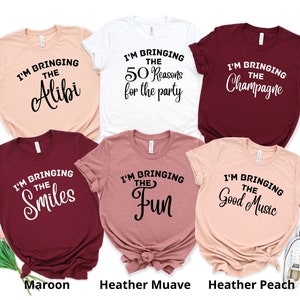 May include: Five t-shirts in different colours with text that reads "I'm bringing the..."  The text is followed by "Alibi", "50 Reasons for the party", "Champagne", "Smiles", and "Fun". The shirts are laid out on a white surface with a pair of white trainers in the bottom right corner.
