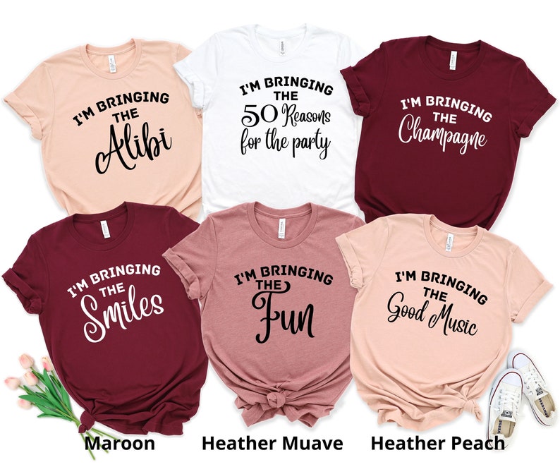 May include: Five t-shirts in different colours with text that reads "I'm bringing the..."  The text is followed by "Alibi", "50 Reasons for the party", "Champagne", "Smiles", and "Fun". The shirts are laid out on a white surface with a pair of white trainers in the bottom right corner.